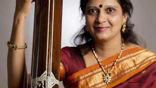 A smiling woman in a red and gold sari holding a tall, wooded, stringed musical instrument.