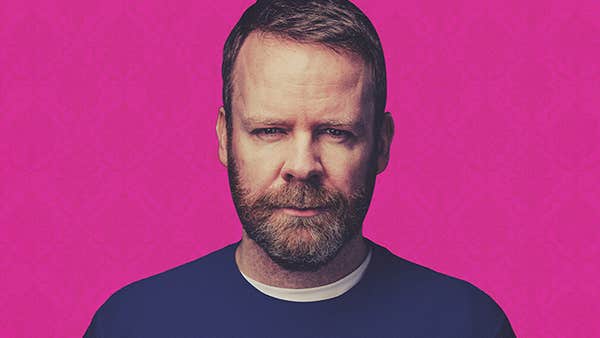 A man with a beard is looking slightly quizzical, in a dark blue jumper, against bright pink background.