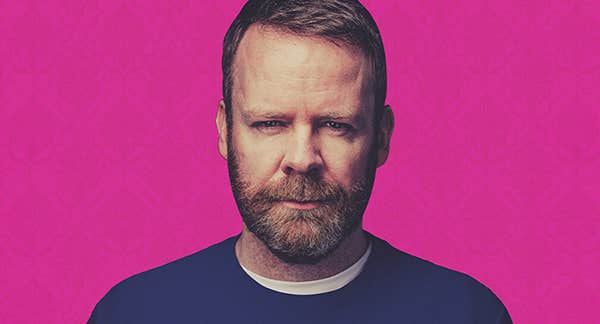 A man with a beard is looking slightly quizzical, in a dark blue jumper, against bright pink background.