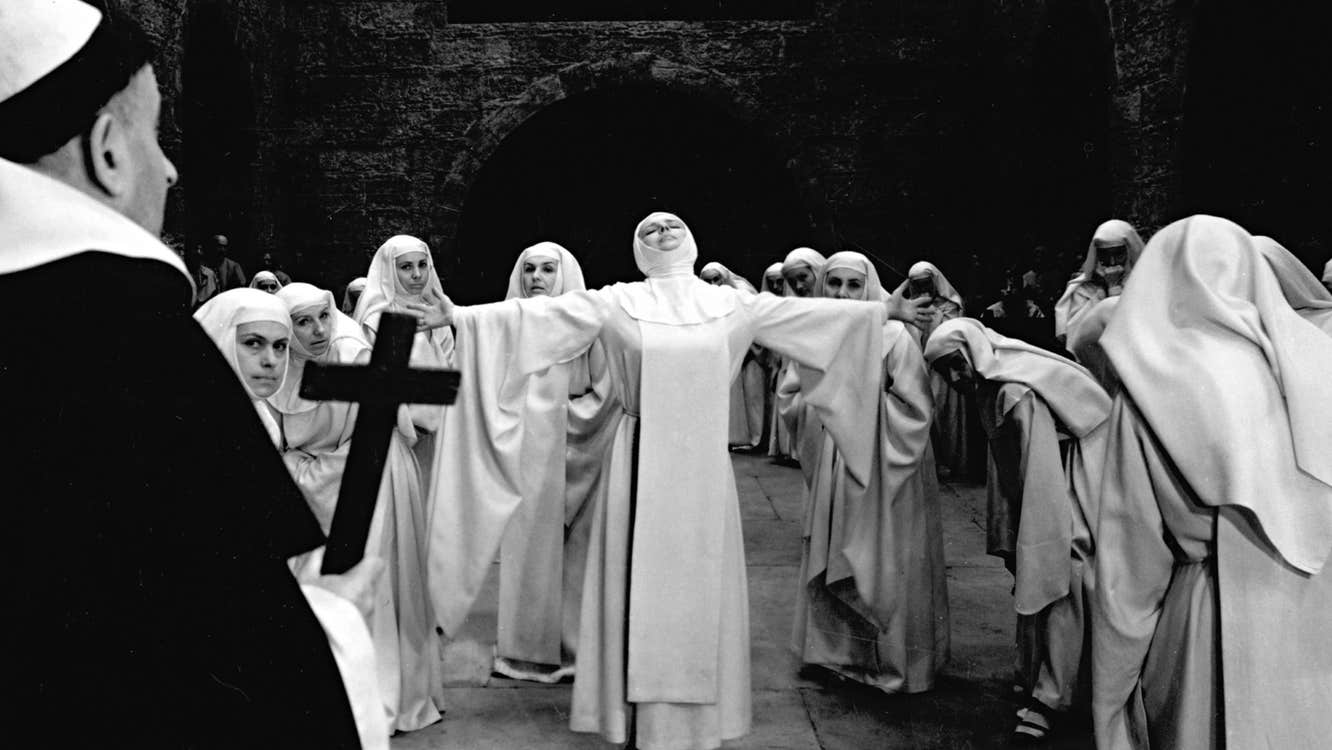 Several nuns in white habits are grouped around one with her arms outstretched.