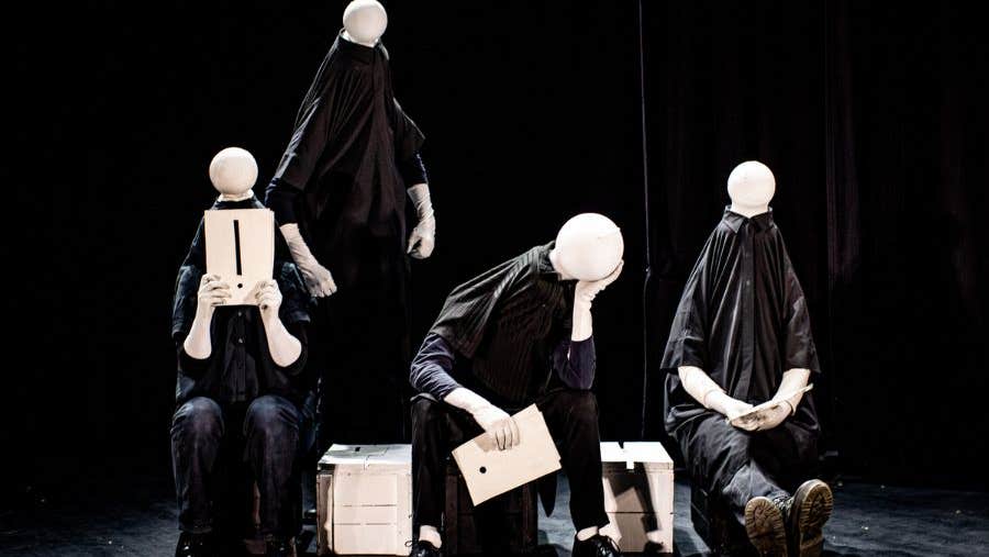 4 plain figures on a stage dressed all in black with plain, small, round blank heads