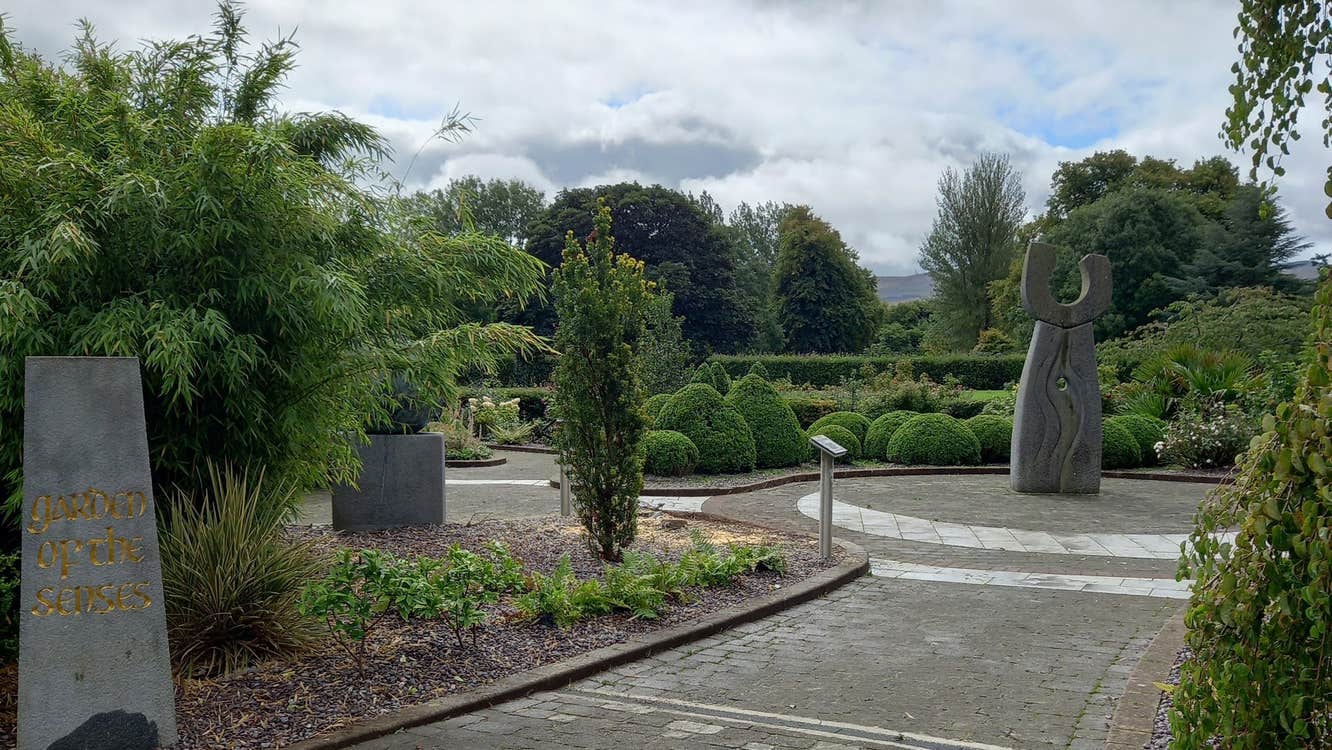 Paved area among trees and shrubbery leading to a tall stone sculpture