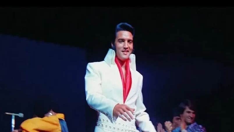 EPiC - Elvis Presley in Concert, slightly blurry photo of smiling man in white suit on black background.
