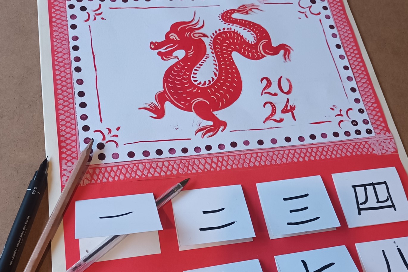 A picture of a Chinese date planner in red and white with a drawn dragon in the centre and 4 square white boxes with simple black lines on across the bottom