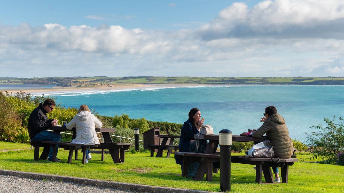 People sitting at picnic tables having lunch with a coastal view in the background