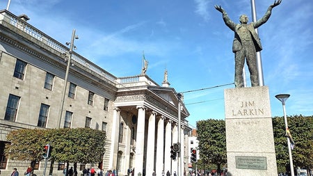 A statue on a plinth of a man standing with arms outstretched beside a busy street