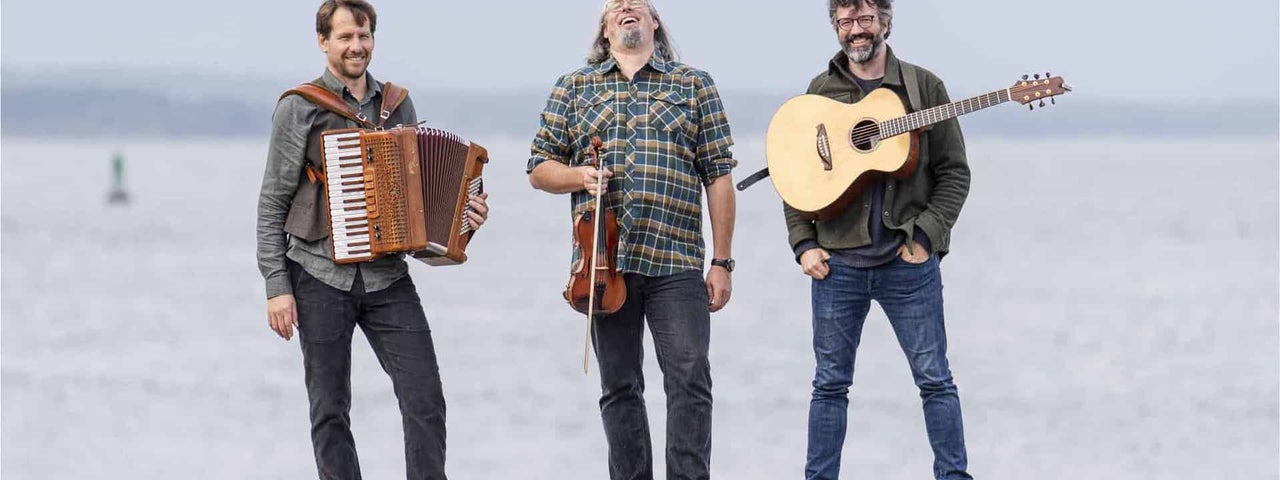 3 men standing on a large rock holding musical instruments with flat, grey sea behind.