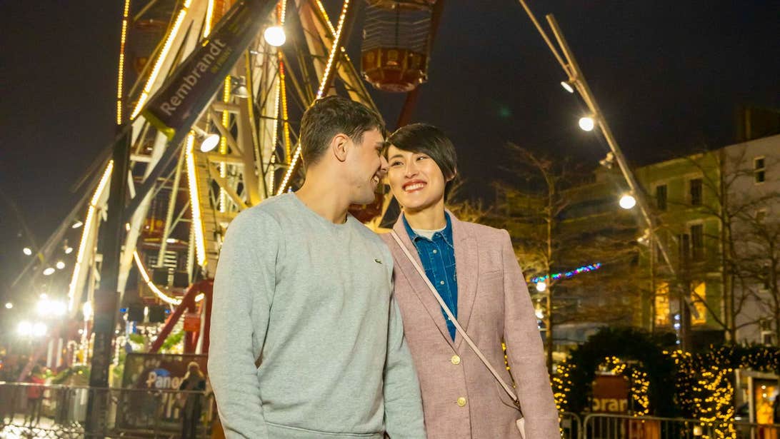 A couple standing infront of the Ferris Wheel that is covered in Christmas lights.