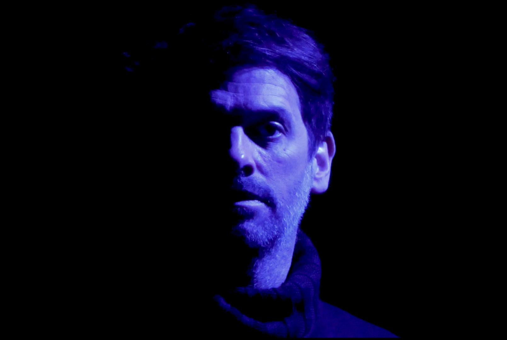 A head view of a man looking serious, dimly lit with blue light against black background.