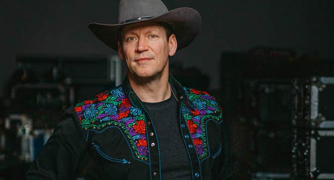 A man in a grey cowboy hat, dark shirt with embroidered flowers in red and green, slightly smiling against dark background.