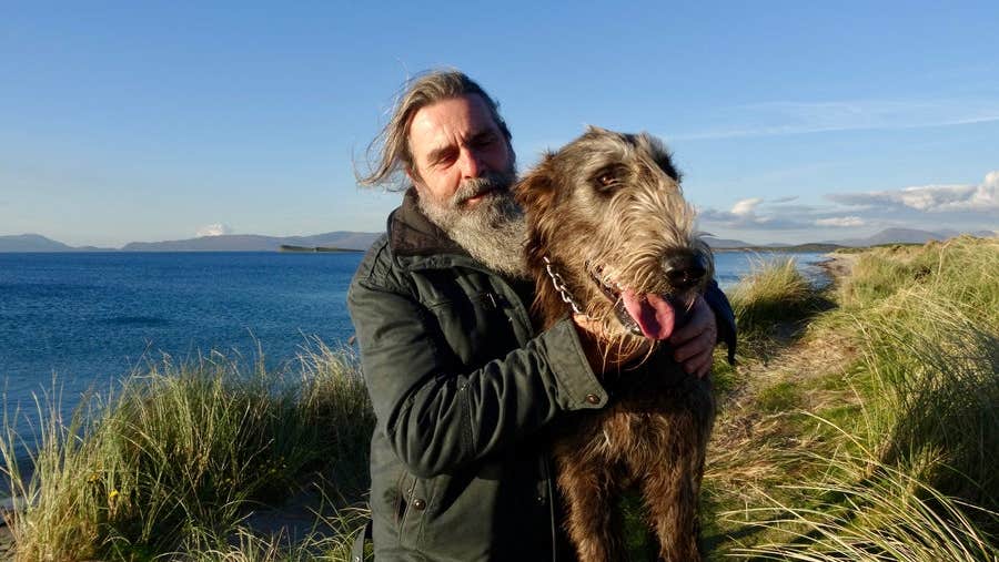 A man posing with an Irish wolfhound in tall grass beside the sea