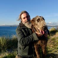 A man posing with an Irish wolfhound in tall grass beside the sea