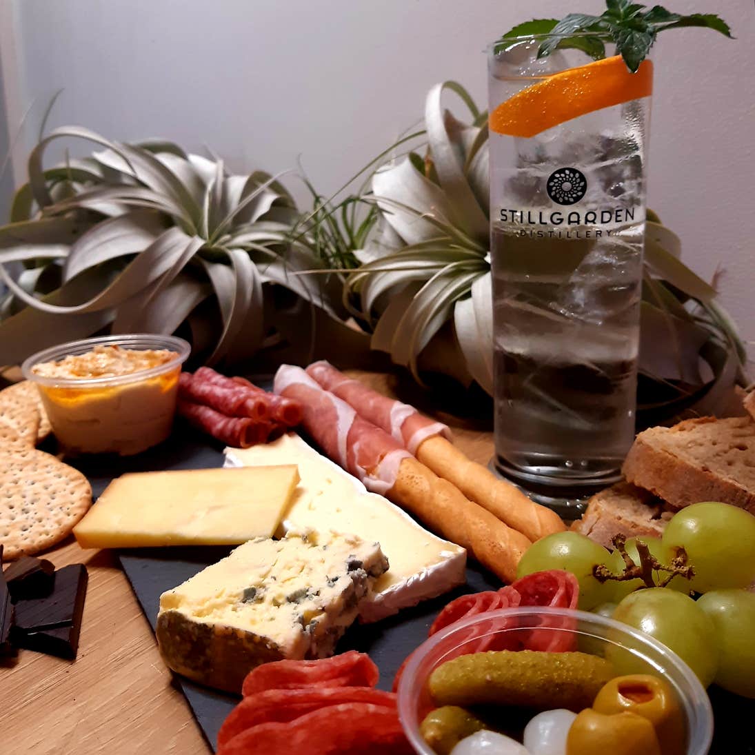 A cheese board on a piece of slate, decorated white various cheeses, cold meats and bread. Served with a glass of Stillgarden gin.