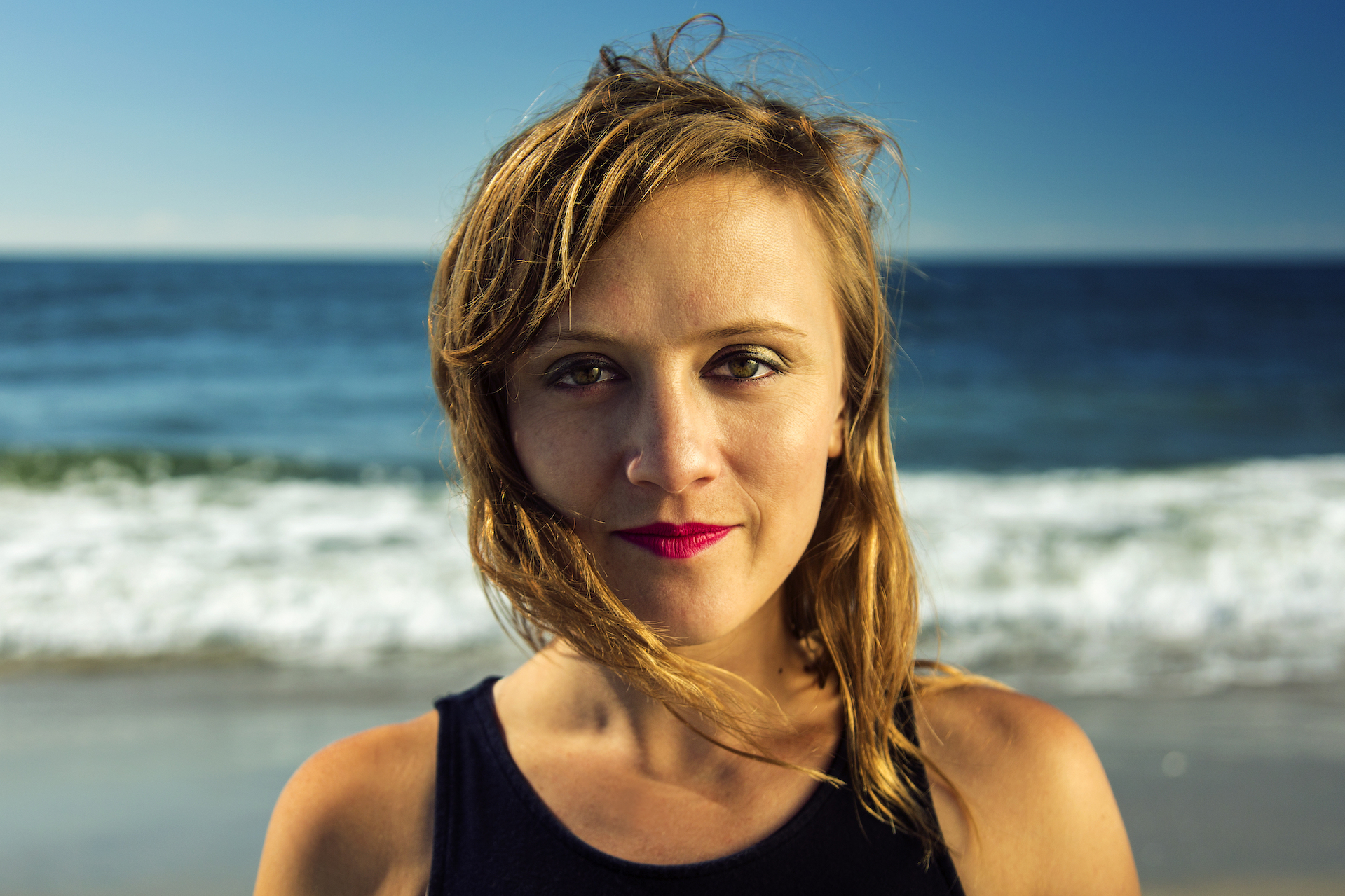 Headshot of a woman looking directly at the camera on a beach with shoreline and sea in the background.