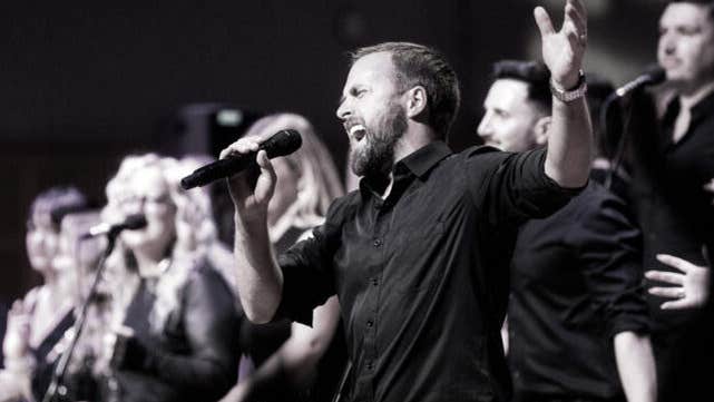 Black and white photo of part of large group singing, with central figure of man singing with emotion holding a mic.