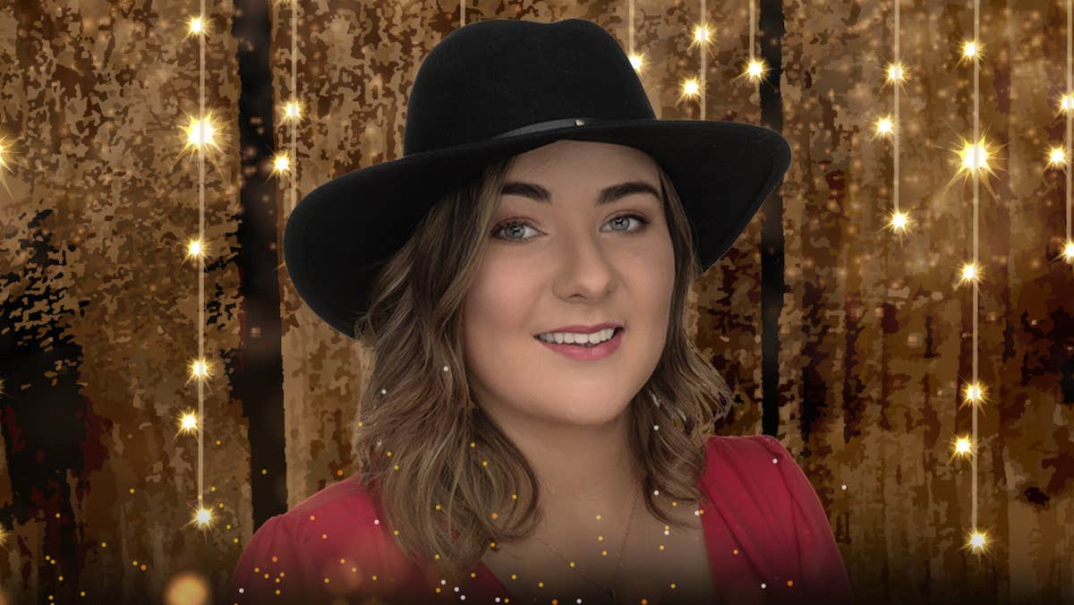 A smiling woman in a black cowboy style hat against dull gold glittery background with hang strings of tiny gold lights.