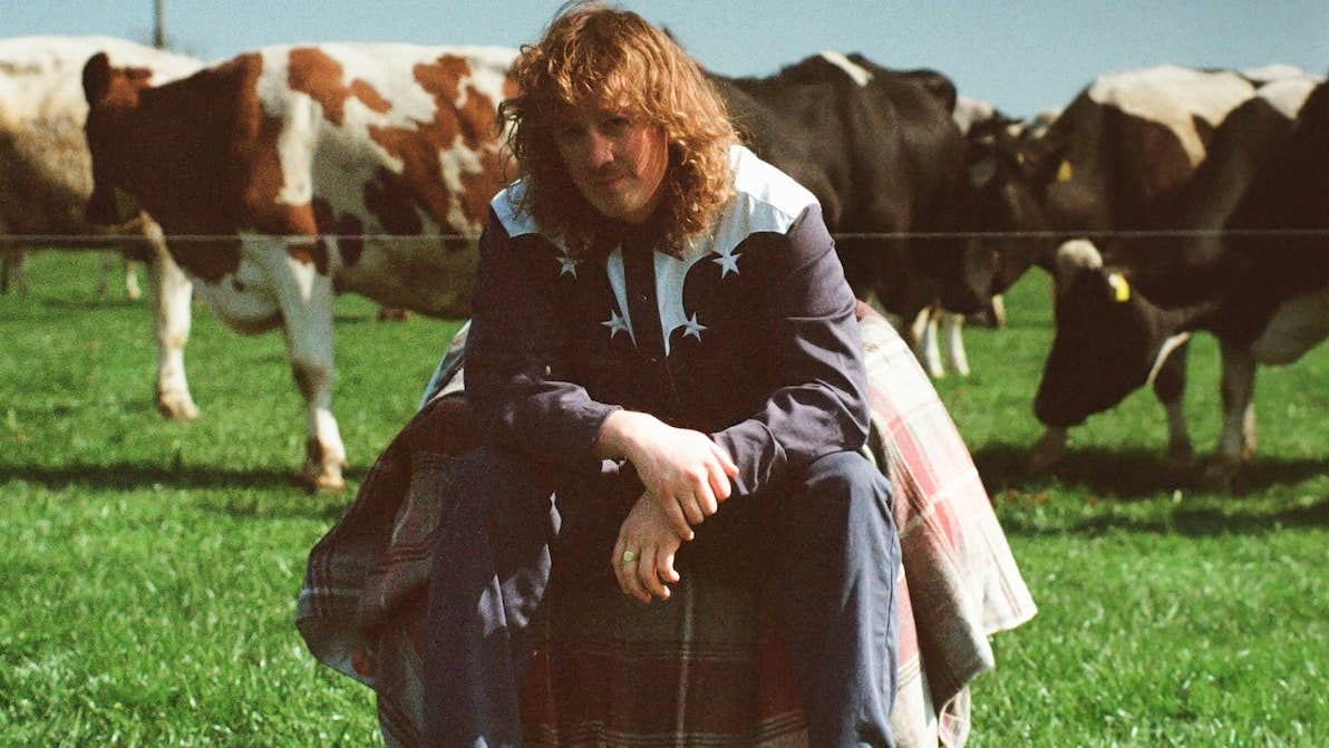 Kean Kavanagh - a man in country style shirt is seated in a field with cows behind.