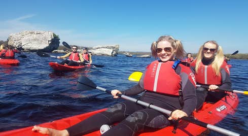 Discover the hidden gems in the Lakes of Killarney with a gentle kayak adventure following a channel that brings you into the depths of the famous Black Valley. 2 women on a red kayak with others in the background.