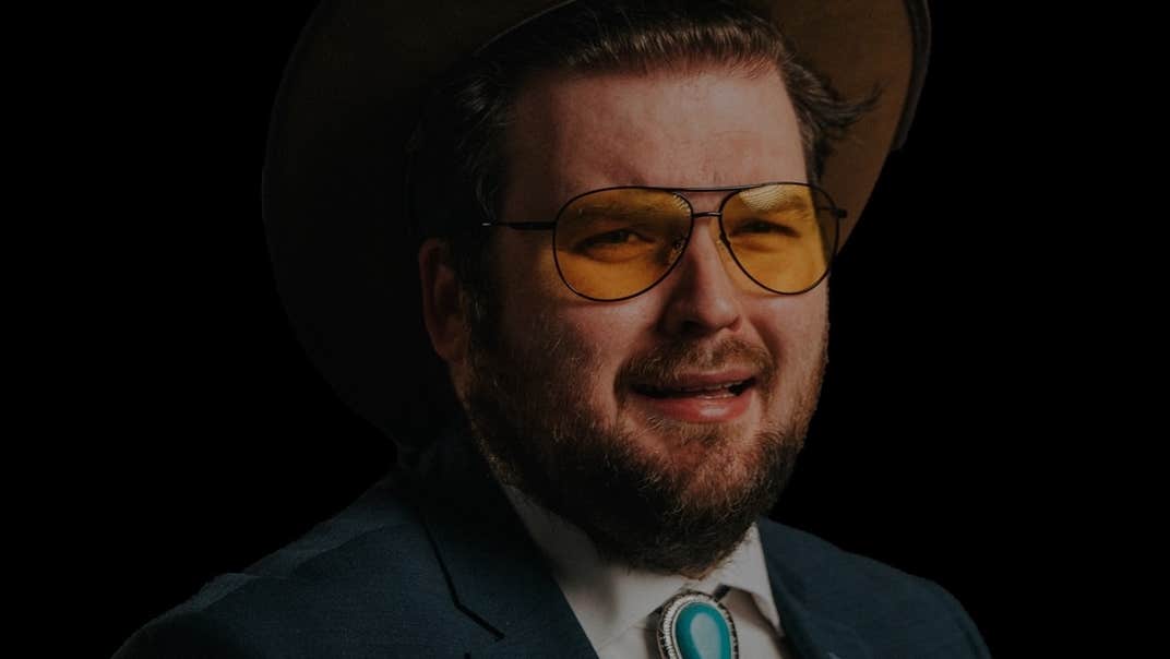 Darkly lit photo of man with beard and yellow tinted glasses in shirt and jacket