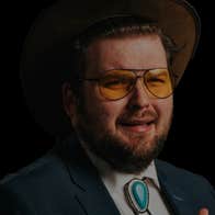 Darkly lit photo of man with beard and yellow tinted glasses in shirt and jacket