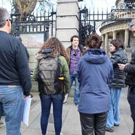A small group of people are standing listening to a man standing outside tall railings and cream stone wall.