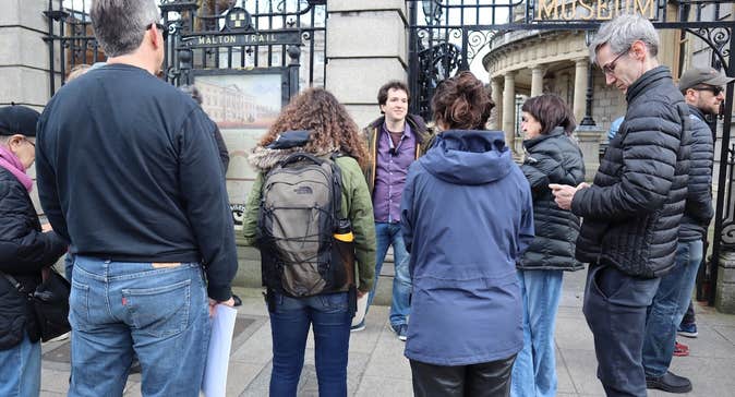 A small group of people are standing listening to a man standing outside tall railings and cream stone wall.