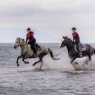 Two people wearing helmets horse riding through water