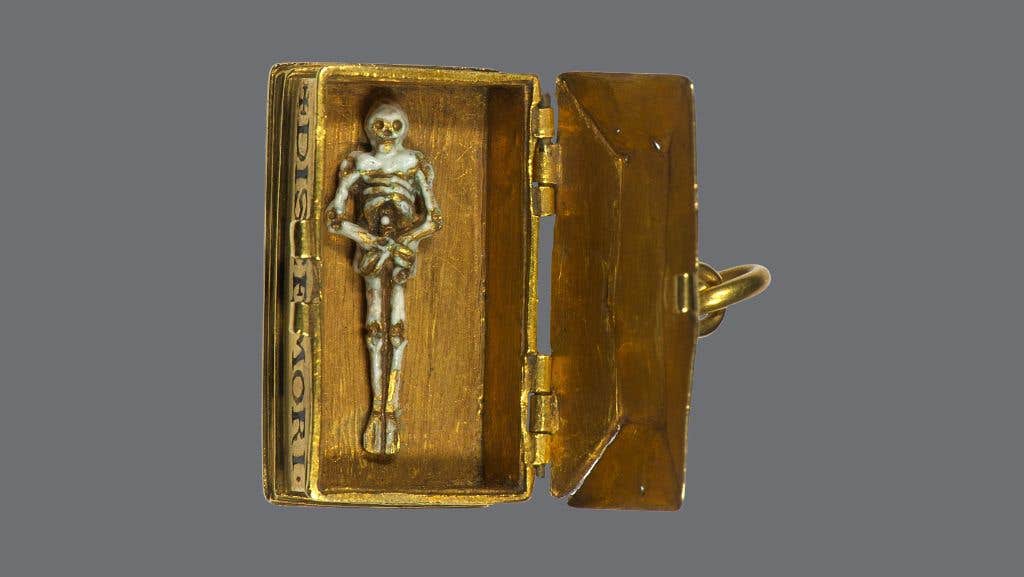 A small, gold coloured ancient box with lid open to reveal a tiny figure of a skeleton lying on the bottom.