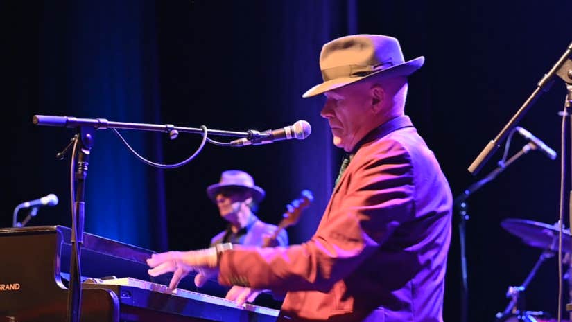 My Leonard Cohen with Stewart D’Arrietta - a side view of a man in a hat with a brim playing a piano on a stage with other musicians.