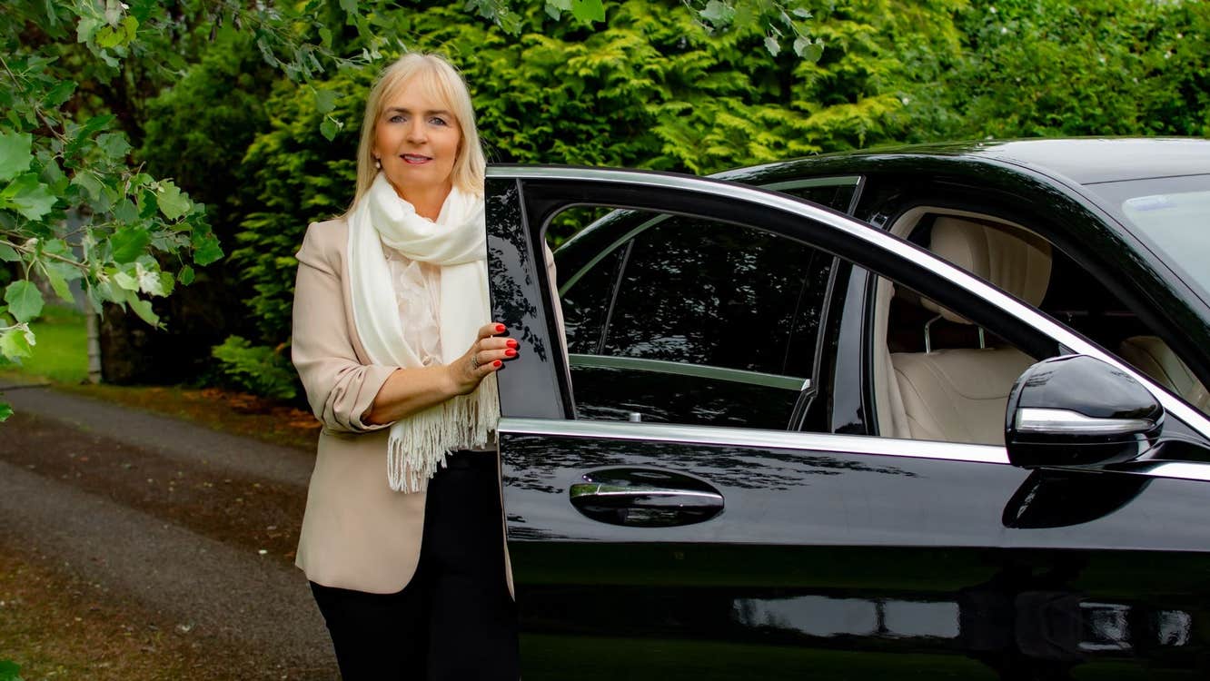 A person standing beside an open car door on a tree lined path