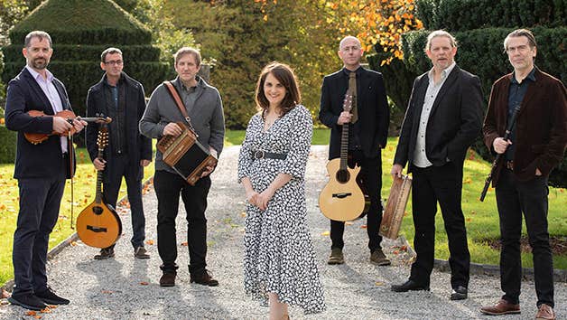 Group of 7 musicians holding instruments standing on a path outdoors in gardens with autumnal tree in background.