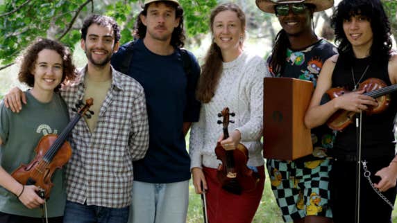 A group of 6 smiling musicians standing under trees