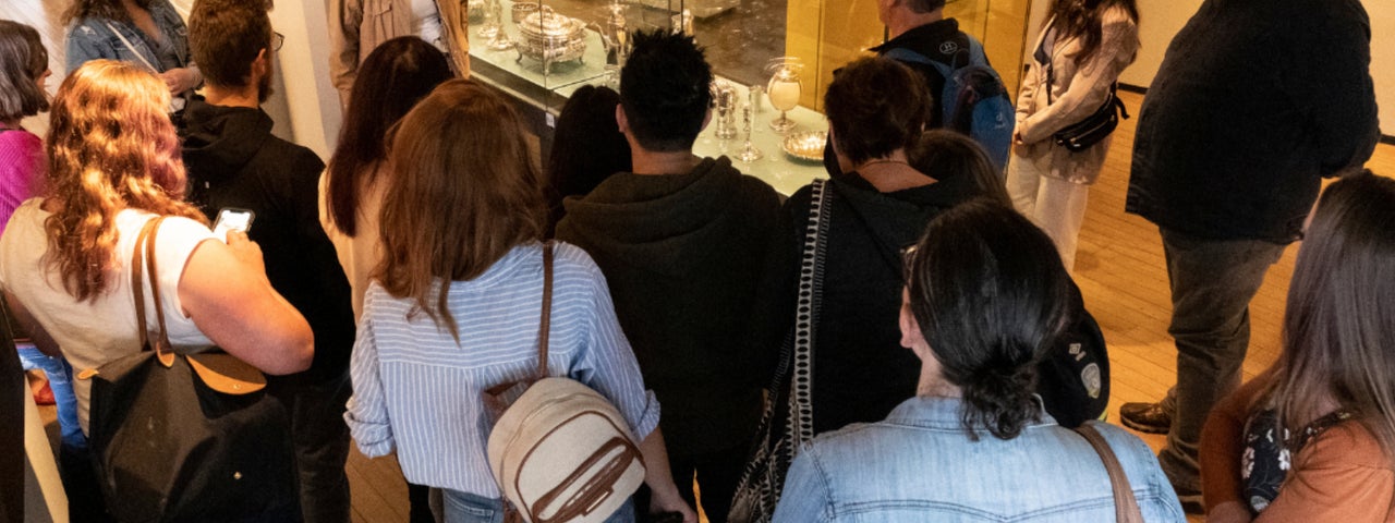 Seen from slightly above, a group of people are gathered around a glass display cabinet with a person standing beside it talking.