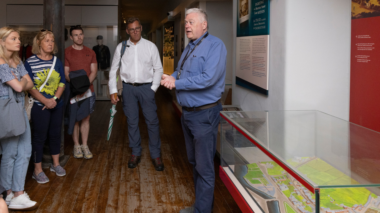 A tour in the Soldiers & Chiefs exhibition. A man is standing in front of a glass cabinet talking to a small group of people.