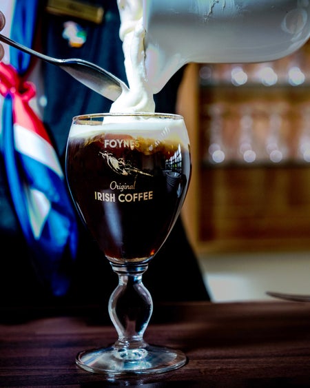 The Irish Coffee Experience, view of a person pouring cream from a jug into a glass with a stem containing a dark coloured drink.
