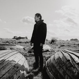Black and white photo of man in dark coat standing between 2 upturned rowing boats looking away into the distance.