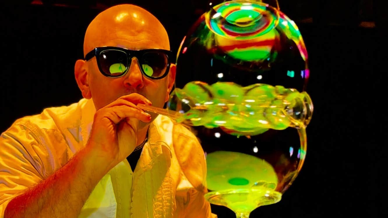 A man in dark glasses is blowing through a small tube with multiple large bubbles at the end