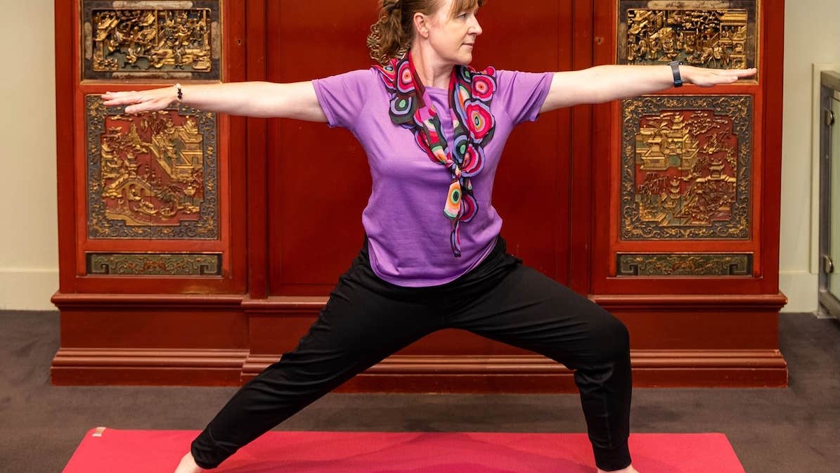 A woman with arms outstretched in a yoga pose with ornate dark red and gold cabinet behind her.