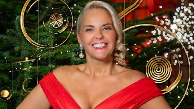 A smiling woman in bright red dress is standing in front of a Christmas tree