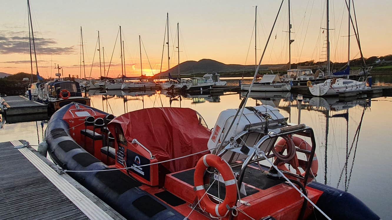 RIB boat docked in the water at a pier at sunset with sail boats and mountain landscape in the background