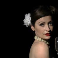 A woman with tied back hair held by white flower is looking over her bare right shoulder at camera with an old style mic in front of her, against black background.