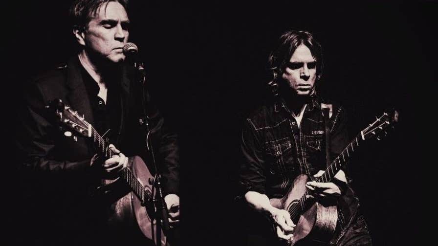 Darkly lit photo of 2 men, both playing guitar, one singing into mic on a stand, against black background.