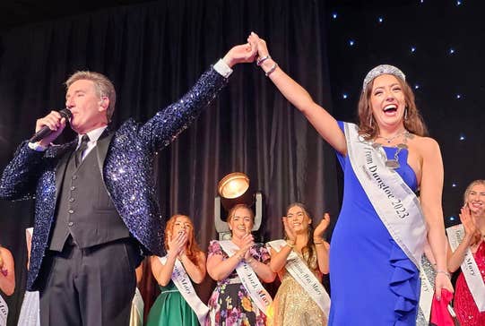 On a stage a man in a dark sparkly jacket, talking into a mic is holding the hand of a woman in a long blue dress, sash and sparkly crown who is laughing, a line of other women in long dresses behind are clapping and smiling