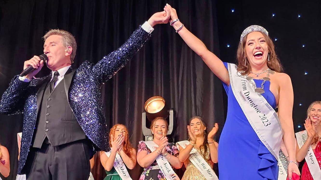 On a stage a man in a dark sparkly jacket, talking into a mic is holding the hand of a woman in a long blue dress, sash and sparkly crown who is laughing, a line of other women in long dresses behind are clapping and smiling