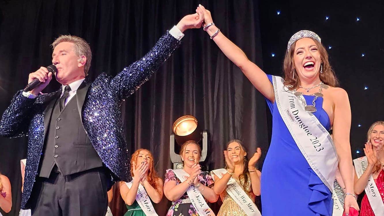 On a stage a man in a dark sparkly jacket, talking into a mic is holding the hand of a woman in a long blue dress, sash and sparkly crown who is laughing, a line of other women in long dresses behind are clapping and smiling