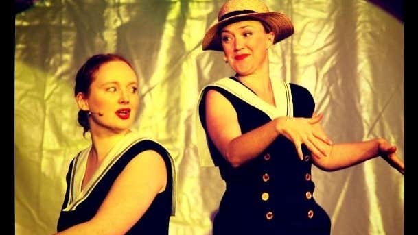 2 women on a stage, on seated at a piano singing, the other dancing beside with gold background.