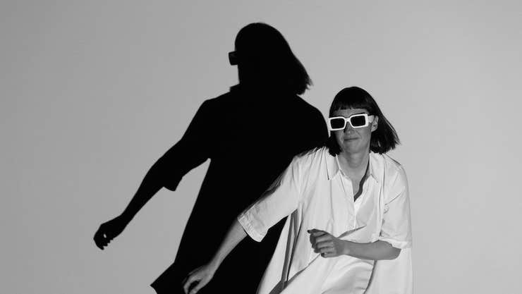 No Woman is an Island by Róisín Stack, a woman seated in dark sunglasses casting a large shadow of herself on plain wall behind.