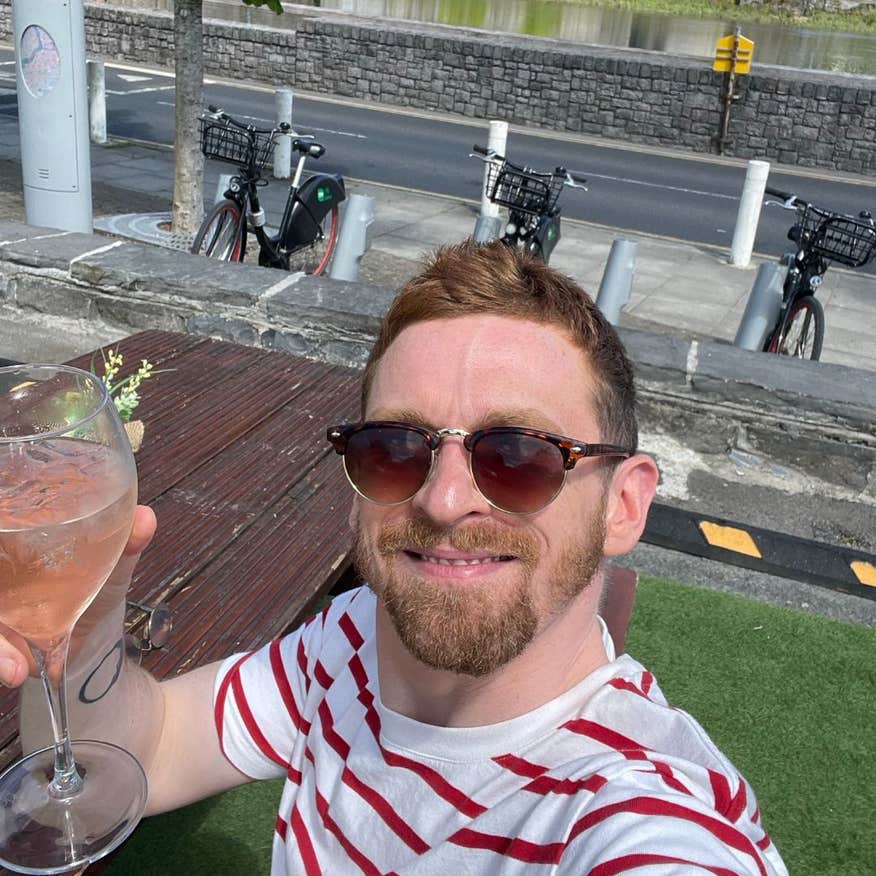 Man in a striped red and white shirt sitting at a bench table with bikes behind him, smiling as he holds up a drink to the camera.