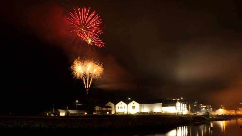 At night looking across water at a white building lit up with red and gold fireworks exploding in the dark sky above.