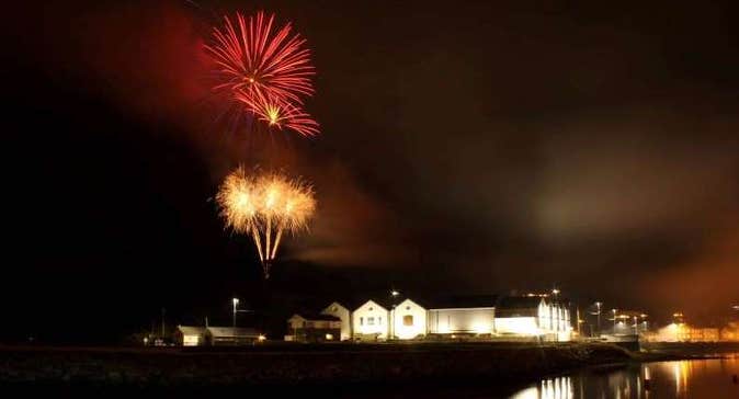 At night looking across water at a white building lit up with red and gold fireworks exploding in the dark sky above.